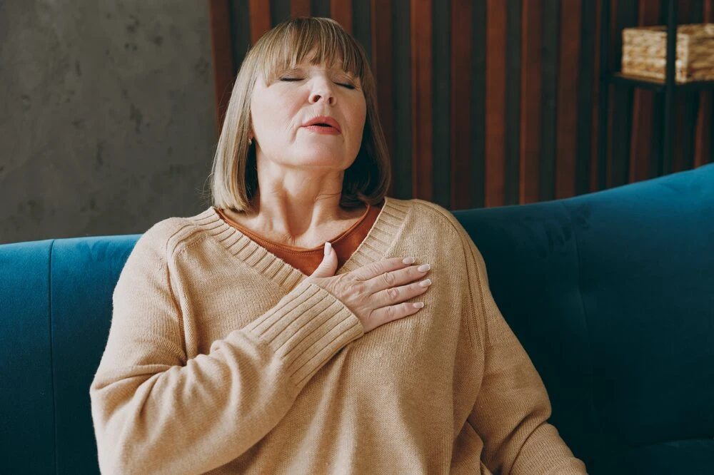 Close up elderly ill sad woman 50s year old wear casual clothes sits on blue sofa put folded hands on heart suffer from pain